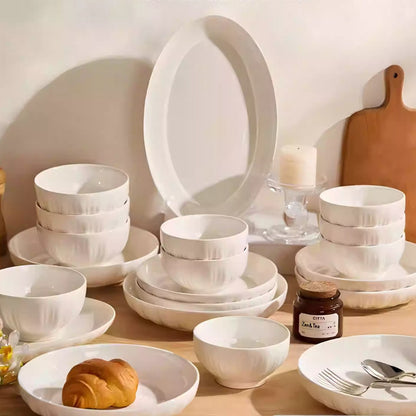 Set of white ceramic dinnerware on a wooden table with a jar of jam and bread.